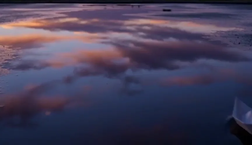 Ein Papierboot treibt in der Dämmerung über eine stille Pfütze, der Himmel spiegelt sich darin